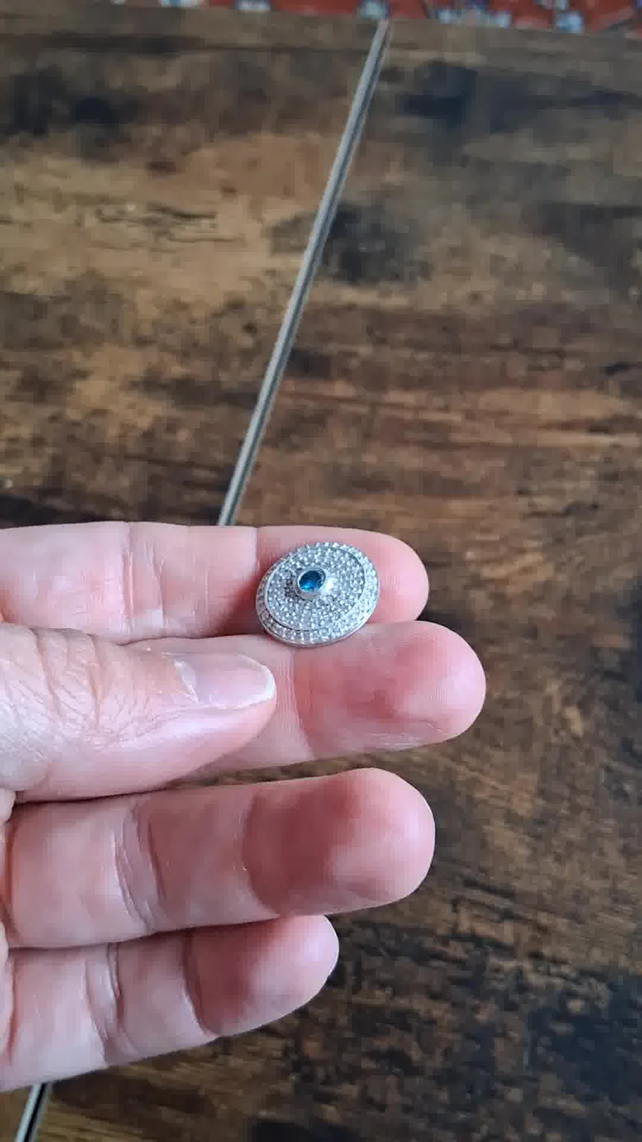 A cast pave-set disc with a blue centre stone, held between two fingers over the bench