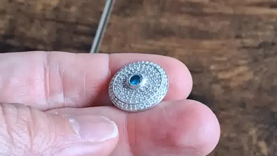 A cast pave-set disc with a blue centre stone, held between two fingers over the bench